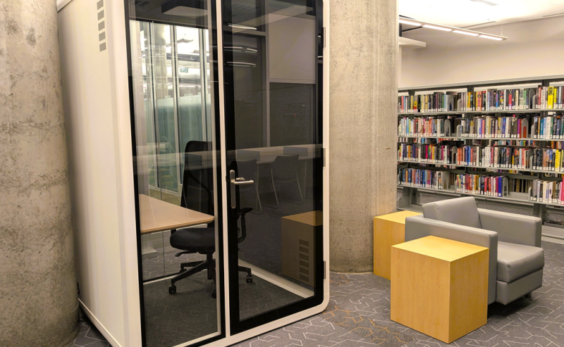 A small, glass-walled privacy booth with a chair and desk inside a modern library space.