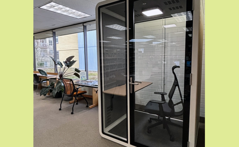 A small, glass-walled privacy booth with a chair and desk inside a modern library space.