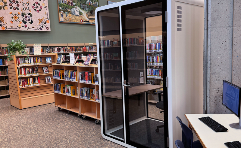 A small, glass-walled privacy booth with a chair and desk inside a modern library space.