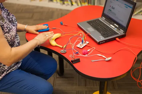 A person plays with a Makey Makey circuit board device by connecting it to various items to create computer commands.