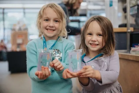 In a library, two young girls smile as they hold their library cards to the camera.