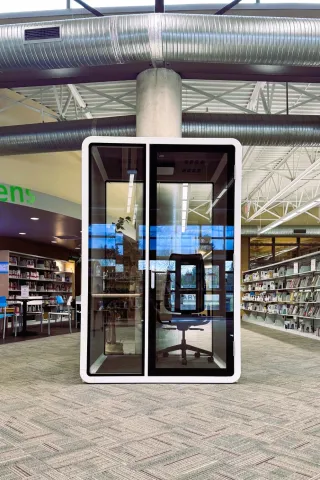 A small, glass-walled privacy booth with a chair and desk inside a modern library space