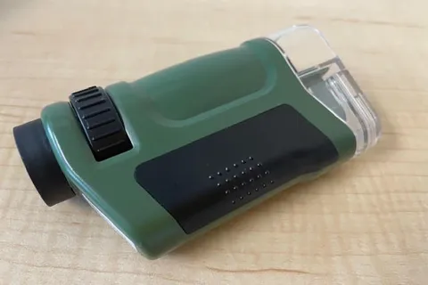 A compact, green monocular microscope with a clear lens, black grip, and focusing wheel, lying on a light wooden surface, conveying practicality.