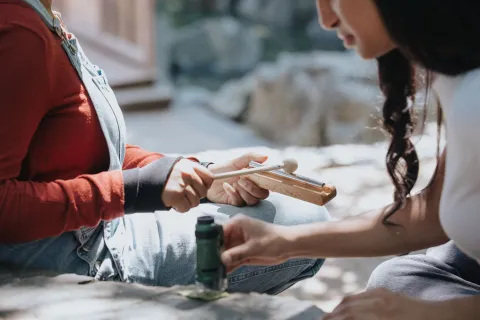 Two teenage girls use components of the FVRL Nature Pack. One uses the chime and mallet while the other views a leaf under the pocket microscope.