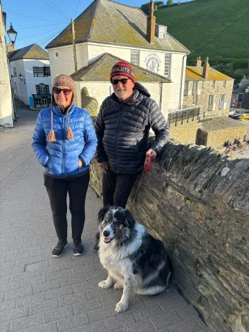Chris Cooper, his wife, and dog Burnett standing near a stone wall on the street in England.
