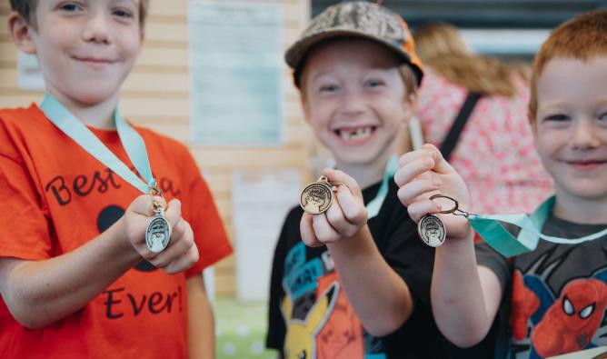 Three kids with smiles on their faces in the library holding up summer reading club medals.