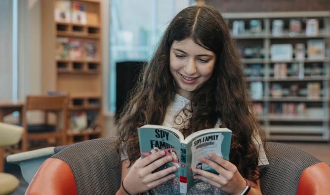 A girl sits in a library, engrossed in reading a book surrounded by shelves filled with various titles.