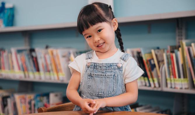 Young Asian girl (5) with braids does crafts at a table with the library book shelves in the background.