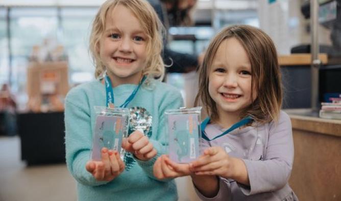 In a library, two young girls smile as they hold their library cards to the camera.