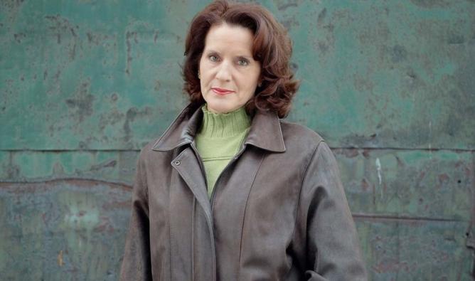 A woman in a brown jacket paired with a green sweater, smiling softly while standing against a neutral background.