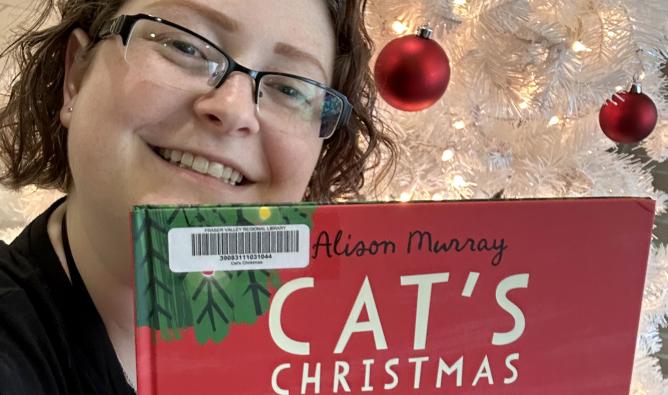 Reading Adviser Sally poses with the picture book Cat's Christmas. A Christmas tree with red ornaments is visible behind her.