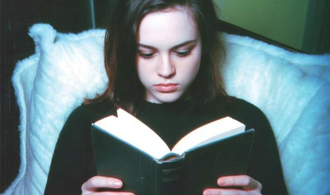 A melancholy teenage girl reclines in bed. She is reading an open book.