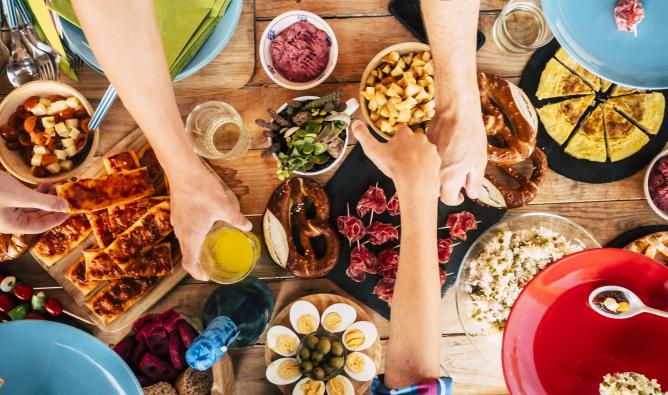 A communal dinner table viewed from above. Dinners reach for various food items including salads, bread, meat, eggs and beverages.