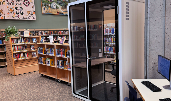 A small, glass-walled privacy booth with a chair and desk inside a modern library space.