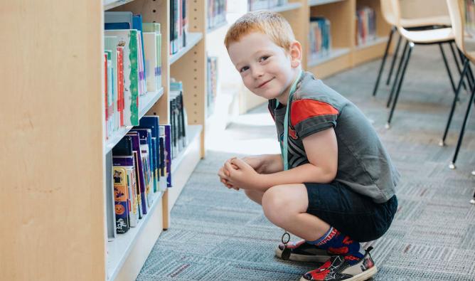 A red-haired boy with Spider-Man shoes squats down to browse a children's bookshelf. He smiles for the camera.