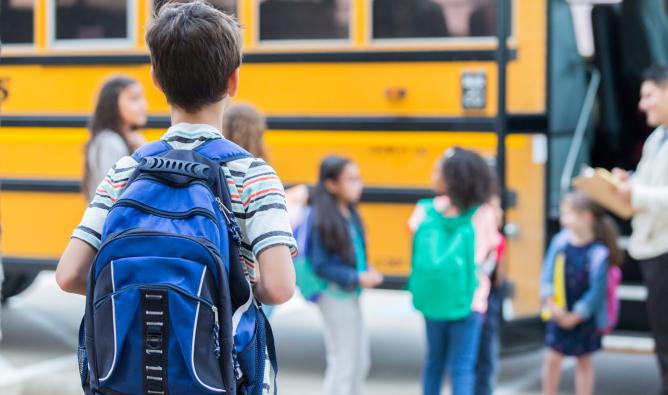 A boy wearing a blue backpack looks towards other school children boarding a yellow school bus.