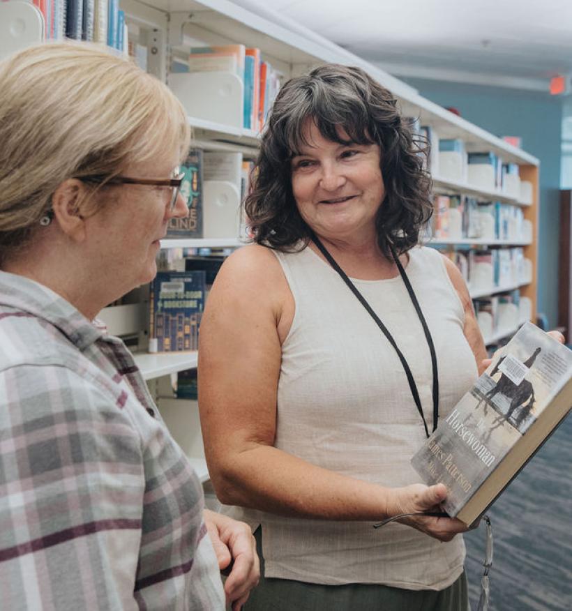 In a library, two women hold books while standing among rows of shelves filled with literature.