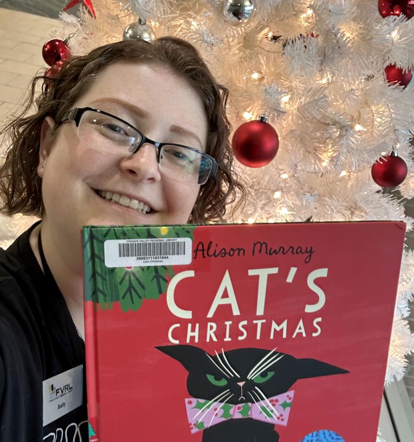 Reading Adviser Sally poses with the picture book Cat's Christmas. A Christmas tree with red ornaments is visible behind her.