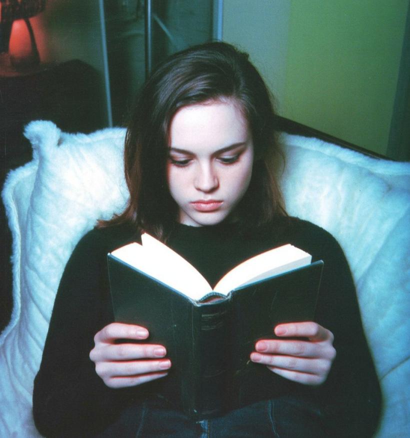 A melancholy teenage girl reclines in bed. She is reading an open book.