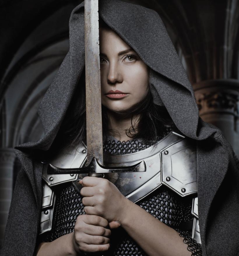 A medieval fantasy image depicts a young woman wearing armour under a grey shroud. She holds a sword in her hands. Gothic arches are visible in the background.
