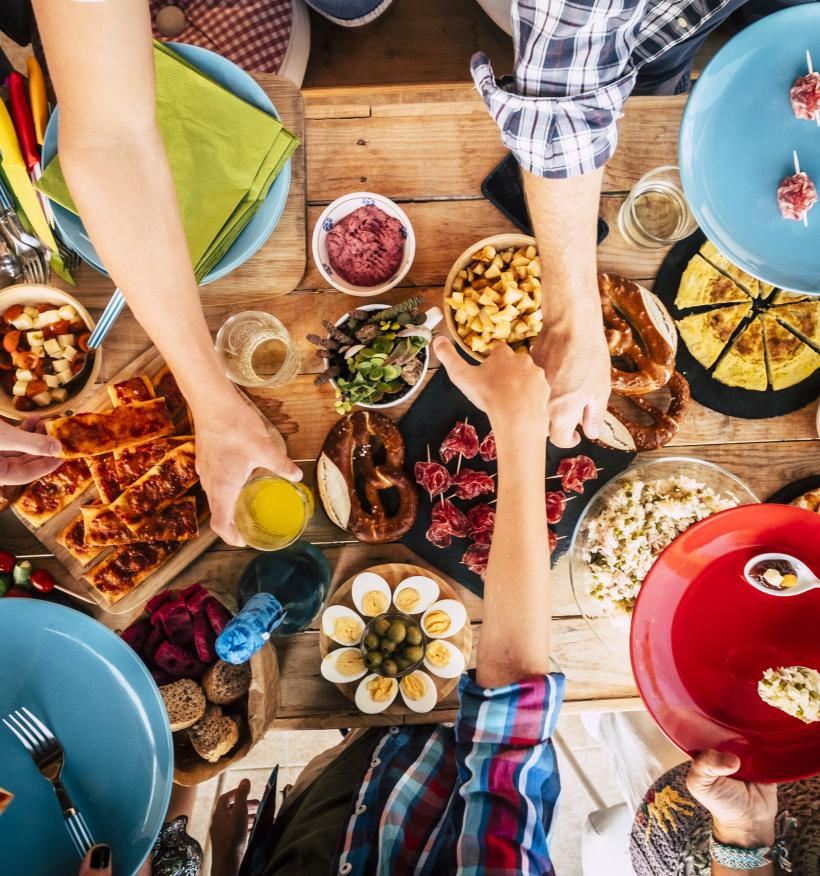 A communal dinner table viewed from above. Dinners reach for various food items including salads, bread, meat, eggs and beverages.