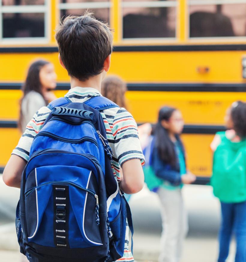 A boy wearing a blue backpack looks towards other school children boarding a yellow school bus.